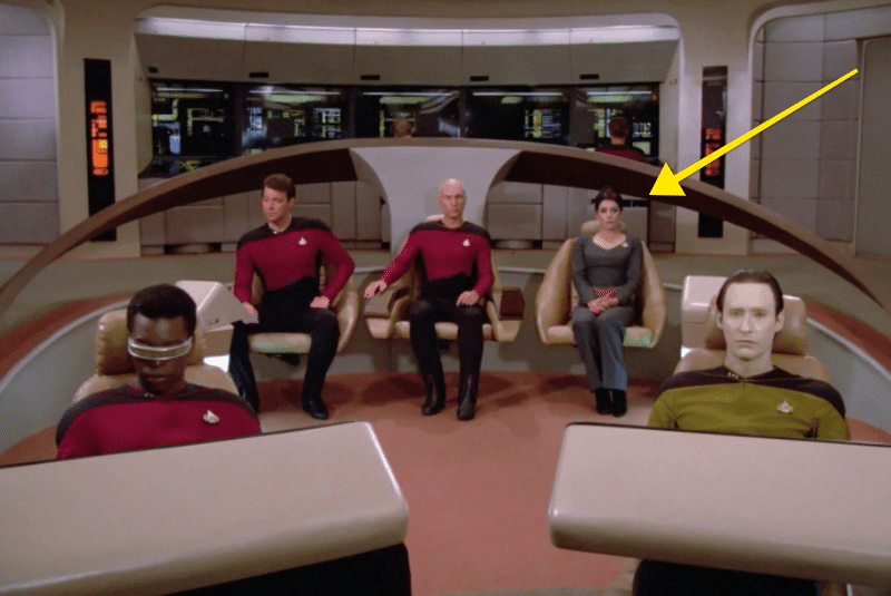 The crew are all sat in the bridge in their seats, with a yellow arrow pointing at Deanna Troi